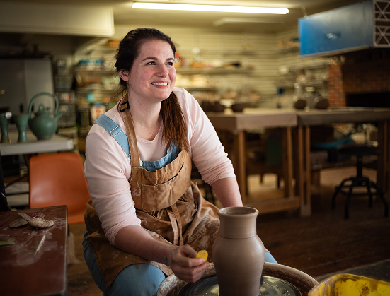 Smiling woman in an apron shaping clay on a pottery wheel in a ceramics studio.