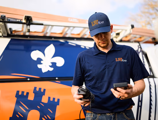Technician in navy LFT Fiber uniform holding cables and device, standing beside a van with a castle and fleur-de-lis logo.