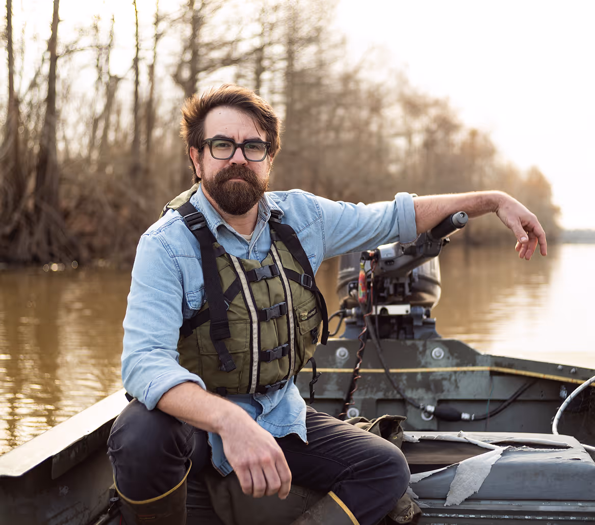 Man with glasses, beard, and life vest sitting in a motorboat on calm water with trees in the background.
