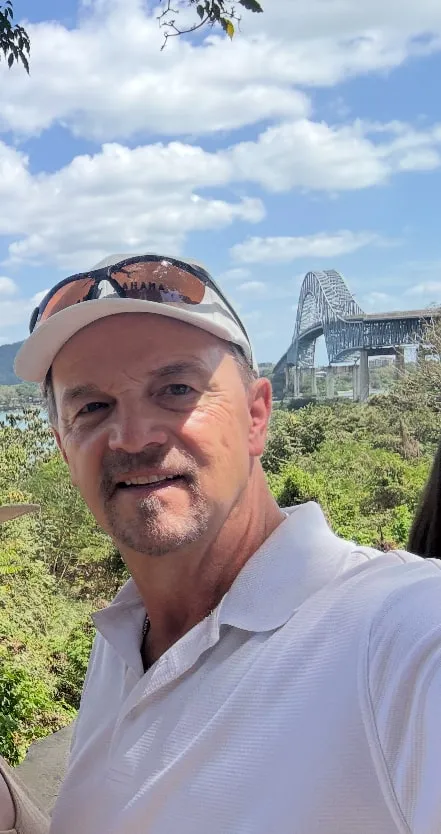 Smiling man in white shirt with bridge and greenery in background