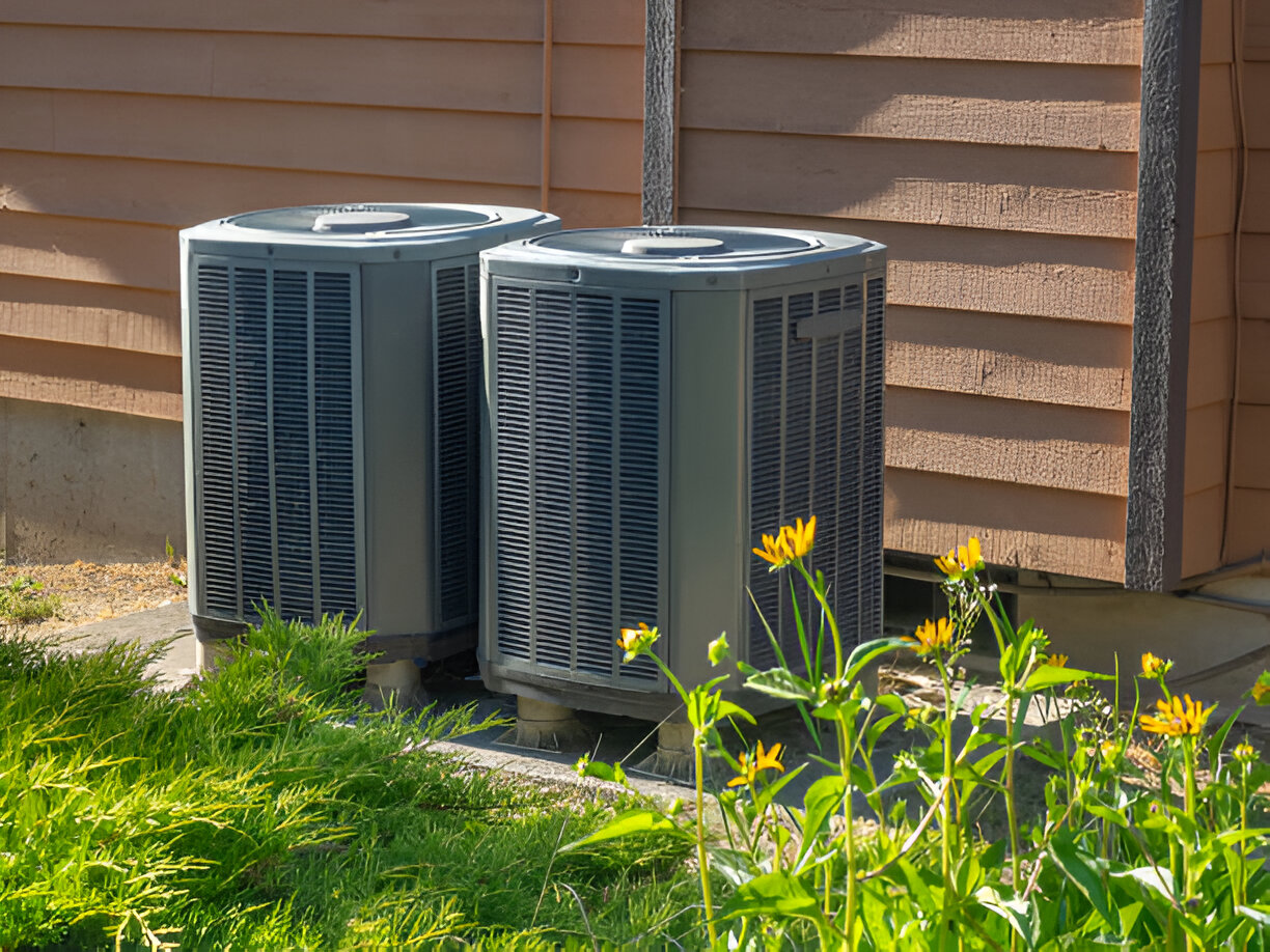 Two air conditioning units next to house with yellow wildflowers