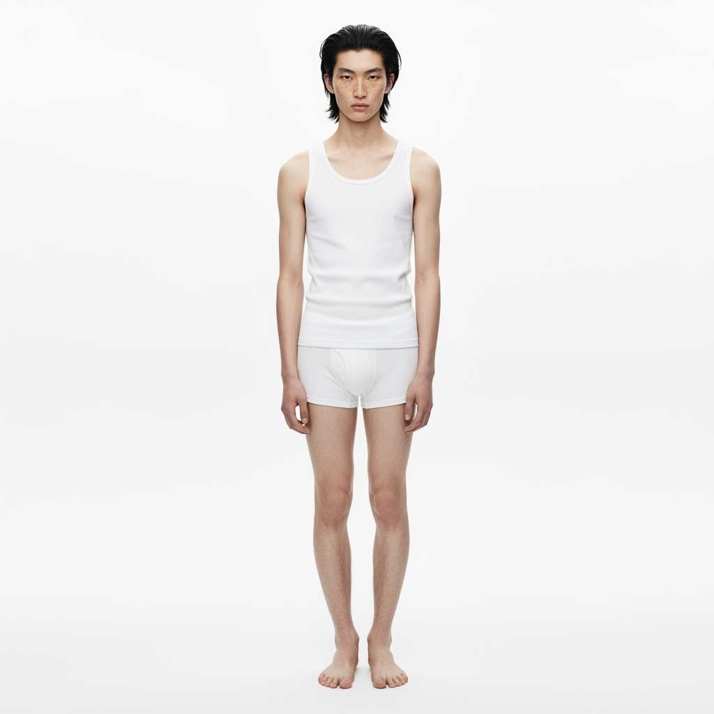 A person with dark, medium-length hair, standing barefoot and wearing a plain white tank top and white boxer briefs against a light gray background. They are looking directly at the camera.