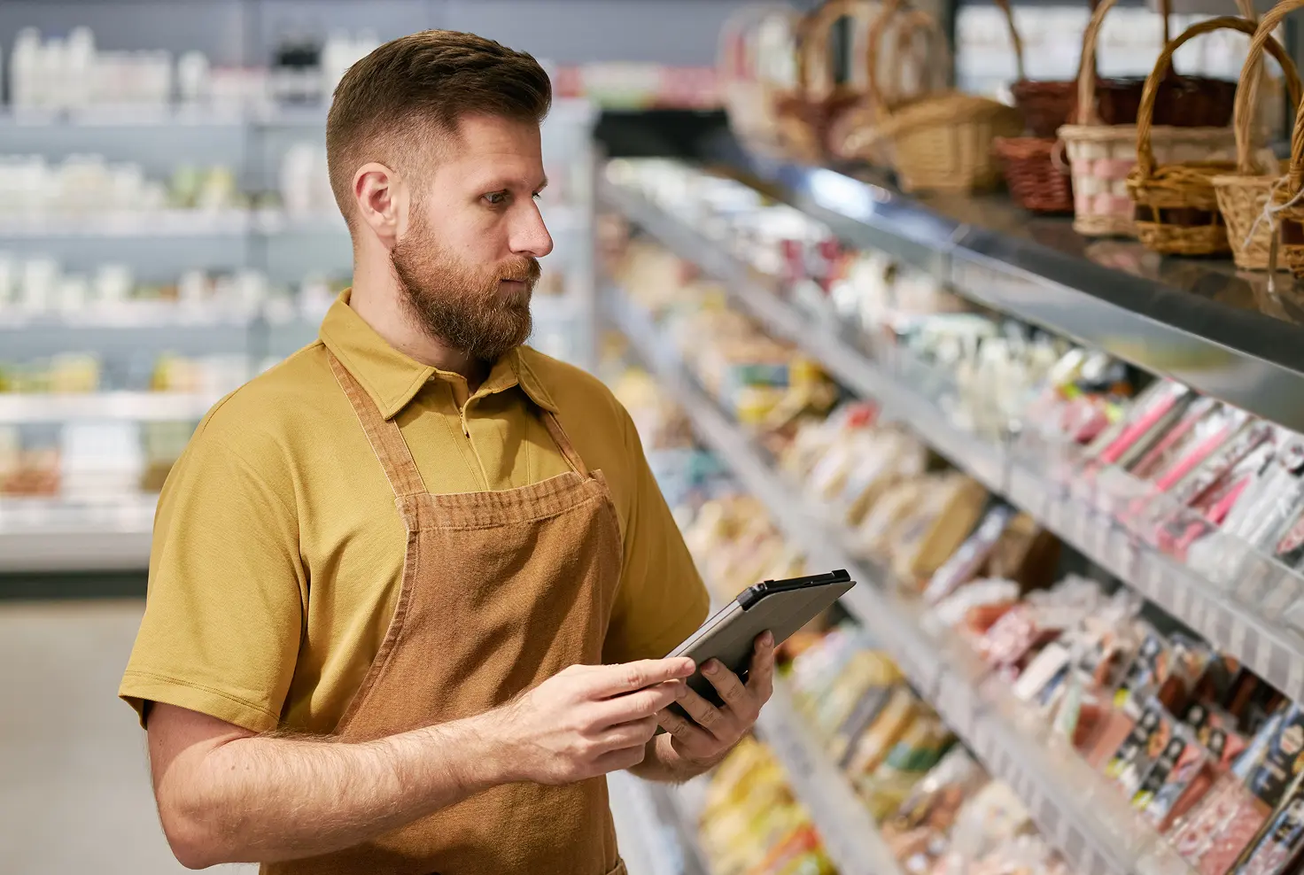 Employé d'épicerie avec tablier utilisant une tablette numérique devant des étagères remplies de produits alimentaires.