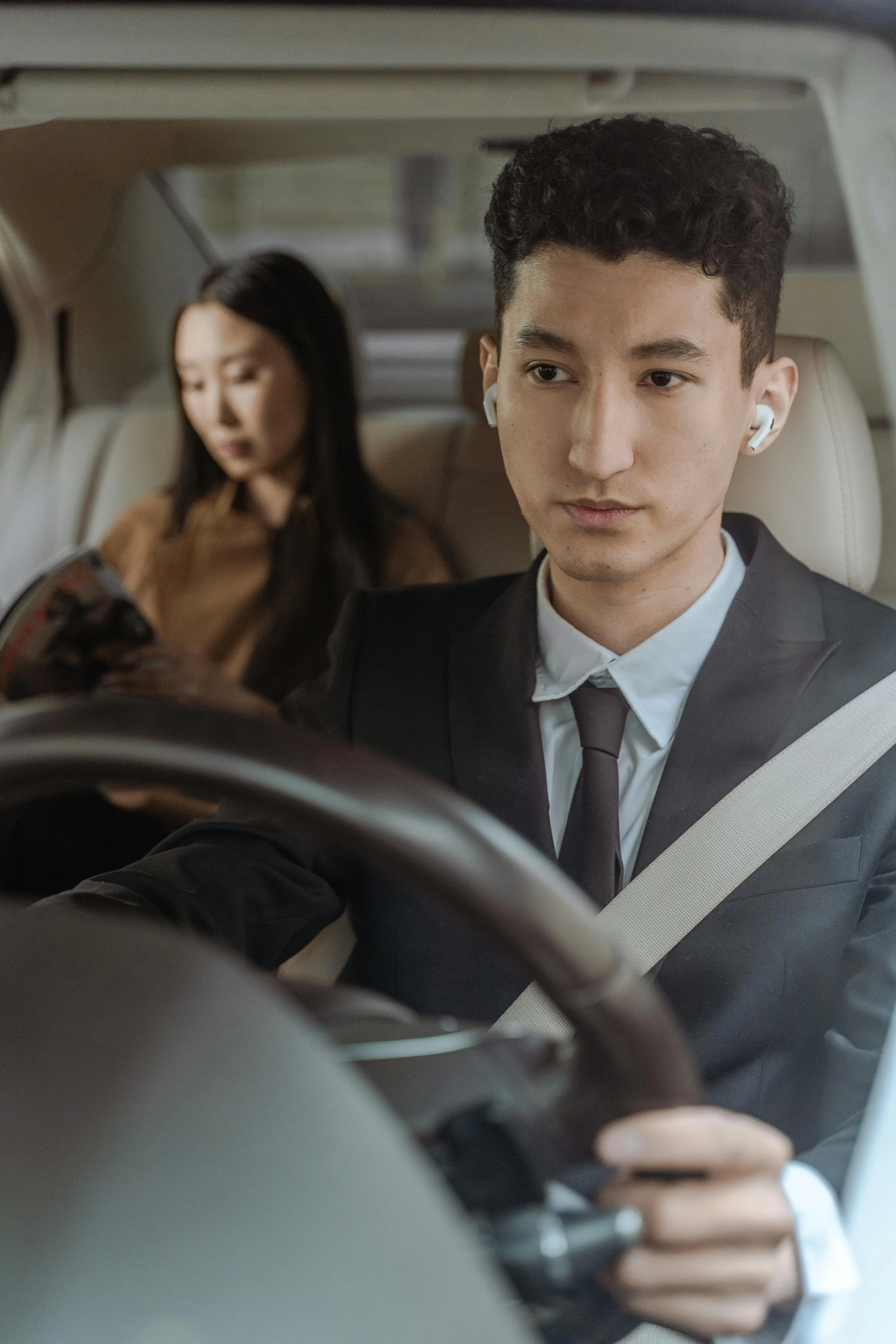 Young man in a suit wearing wireless earbuds and driving a car with a woman reading a magazine in the back seat.