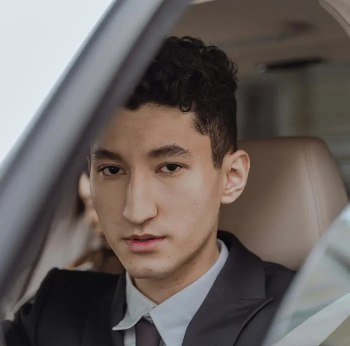 Close-up of a young man in a suit sitting in a car, looking confidently at the camera.