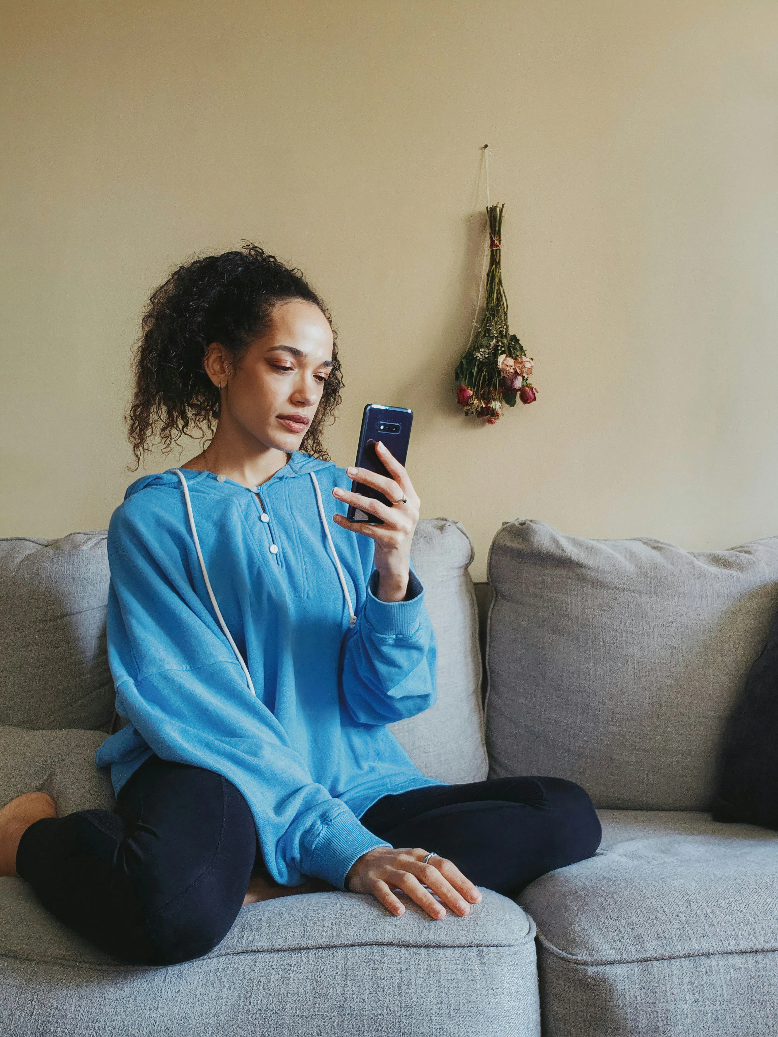 Young woman in a blue hoodie sitting cross-legged on a gray couch, looking at her phone.