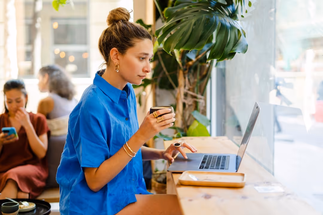 Woman in a blue shirt working on a laptop in a bright cafe.