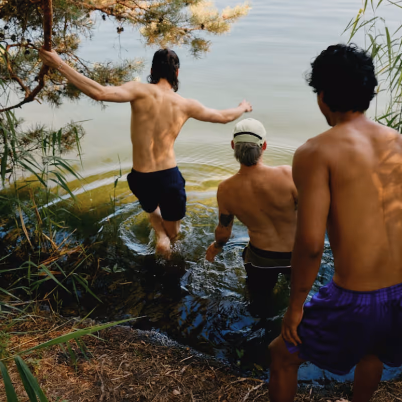 Three men from behind entering a lake or river from a grassy bank.