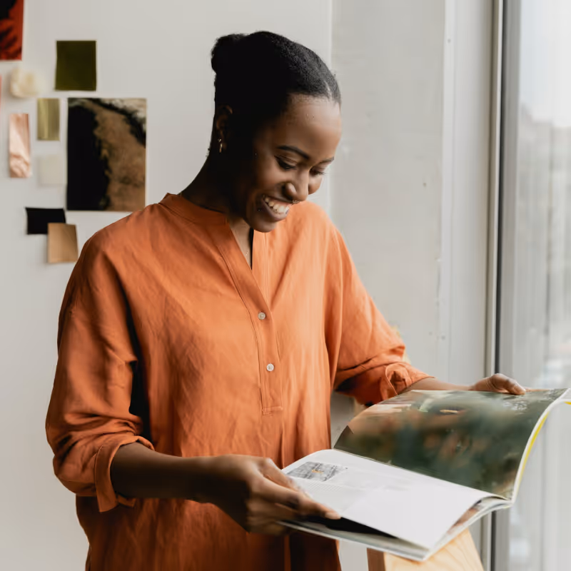 Smiling woman in an orange shirt reading a large magazine by a window, with a fabric mood board behind her.