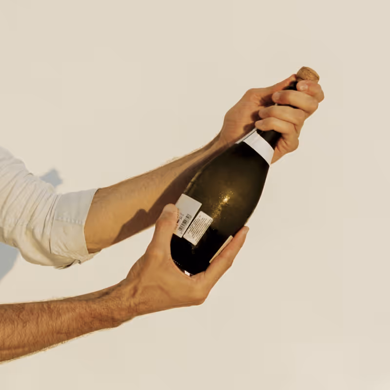 Hands of a person in a white shirt opening a dark bottle of champagne or sparkling wine.