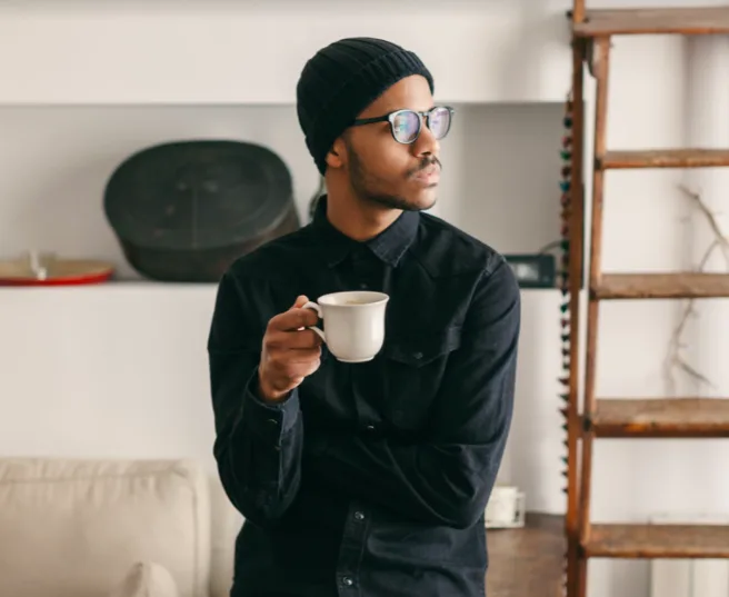 Man wearing glasses and a black beanie holding a white coffee cup looking to the side in a cozy indoor space.