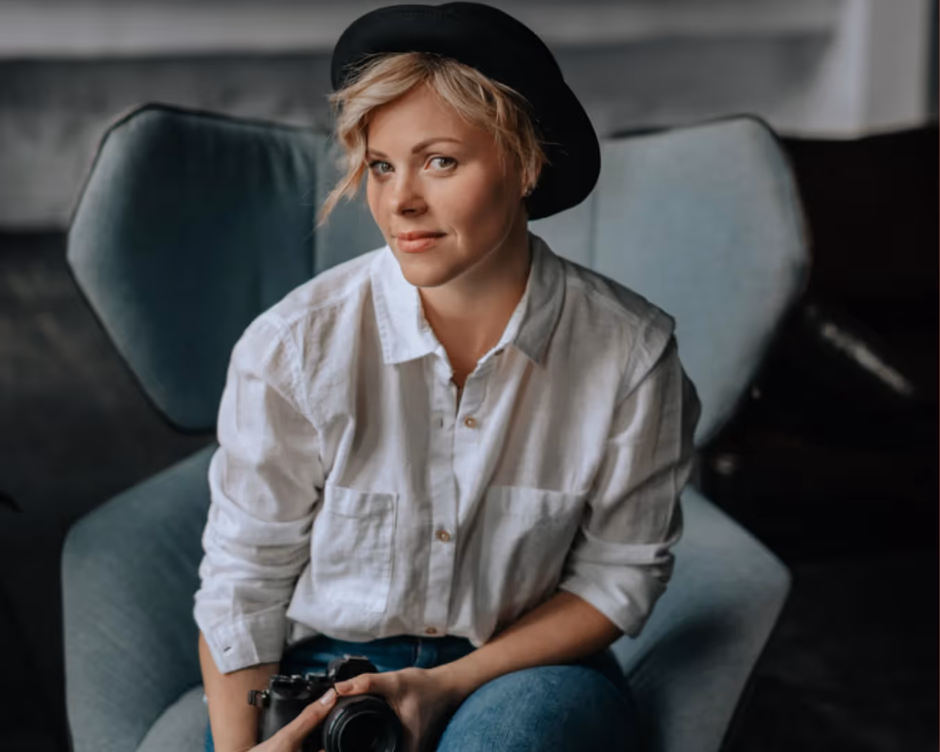 Smiling woman wearing a black hat and white shirt sitting in a gray armchair holding a camera.