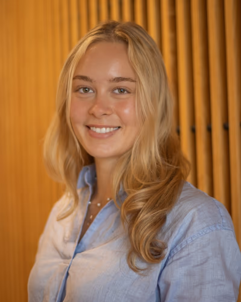 Young woman with blonde hair smiling in light blue shirt against gold curtain background.