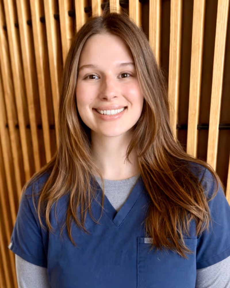 Smiling young woman with brown hair wearing blue scrubs and gray sleeve shirt
