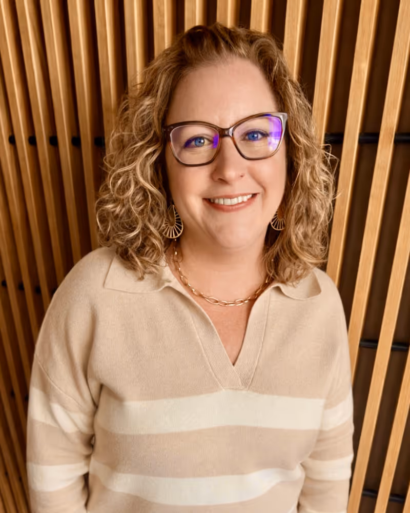 Woman with curly auburn hair wearing glasses and beige striped sweater smiling at camera