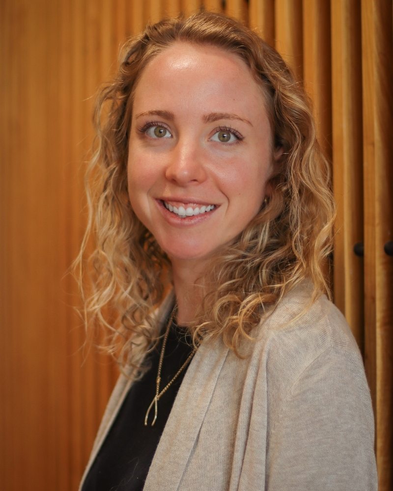 Smiling woman with curly reddish-brown hair wearing beige cardigan against orange backdrop