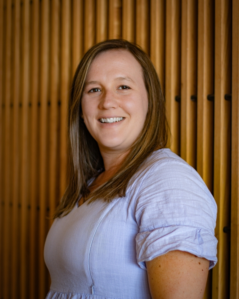 Woman with brown hair smiling at camera wearing light blue shirt against gold curtains