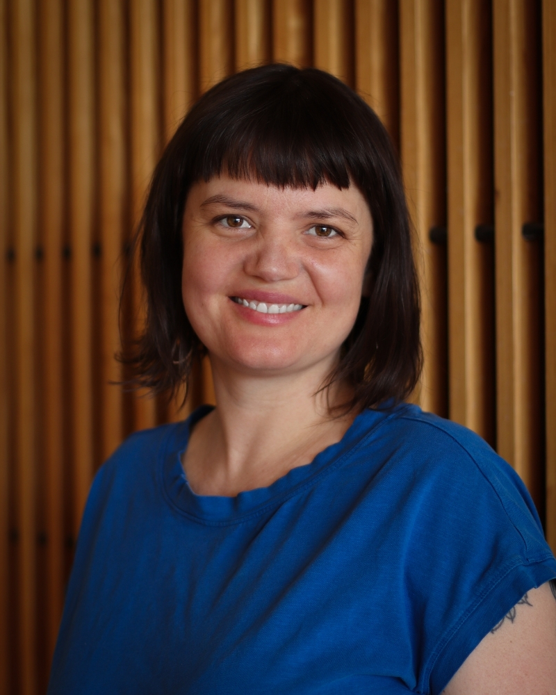 Woman with black bangs wearing blue shirt, smiling against striped gold background
