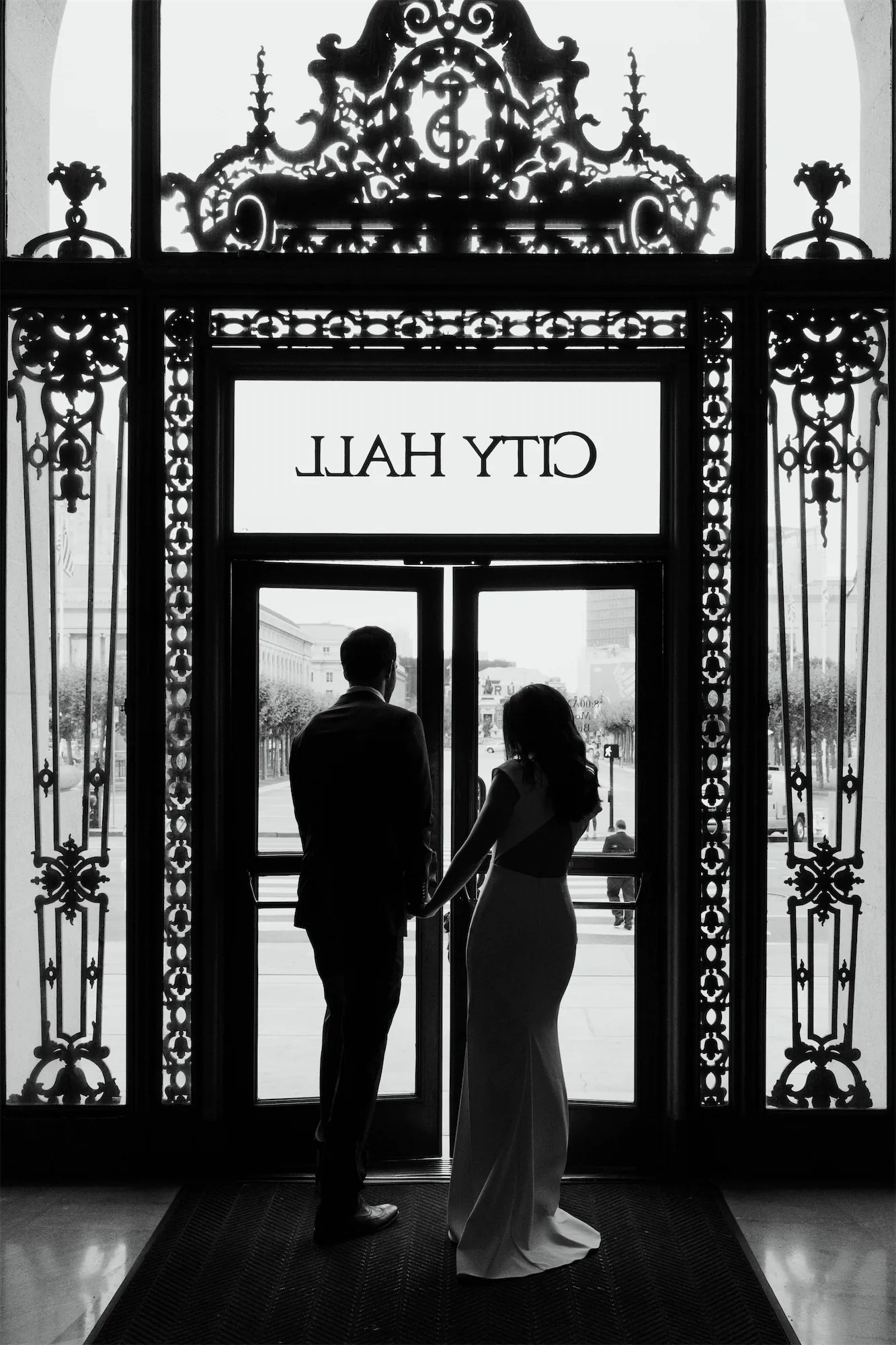 Bride and groom silhouetted hand-in-hand before the ornate iron doors of San Francisco City Hall, black and white film-inspired elopement photography