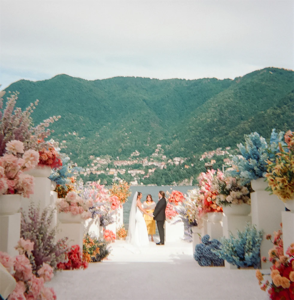 Lake Como wedding ceremony with colorful rainbow floral installation overlooking the lake and mountains in Italy, photographed on film with an editorial, romantic style.