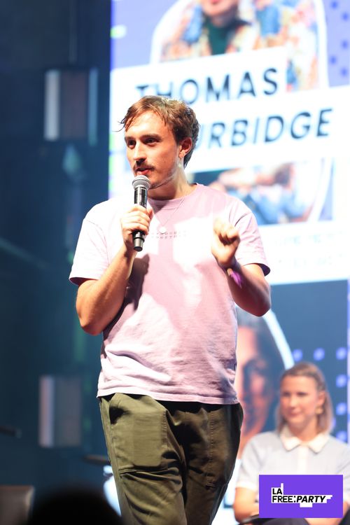 Un homme debout sur scène parlant dans un microphone, portant un T-shirt rose clair et un pantalon olive, avec une femme assise à l'arrière-plan et un grand écran affichant le nom "Thomas Burbidge".