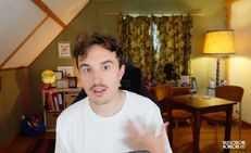 Un homme aux cheveux courts et moustache parle devant une caméra dans une pièce chaleureuse avec une lampe, une table en bois, des livres sur une étagère, et un rideau fleuri à la fenêtre.
