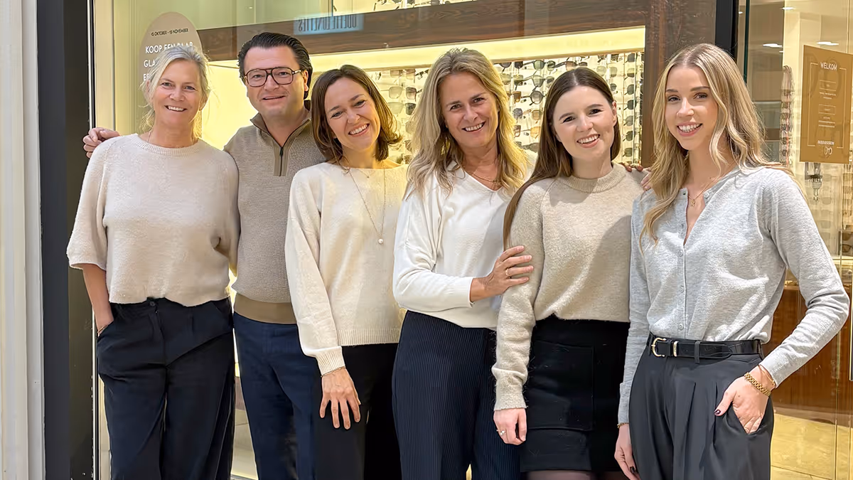 Zes lachende volwassenen poseren samen voor een etalage met brillen.