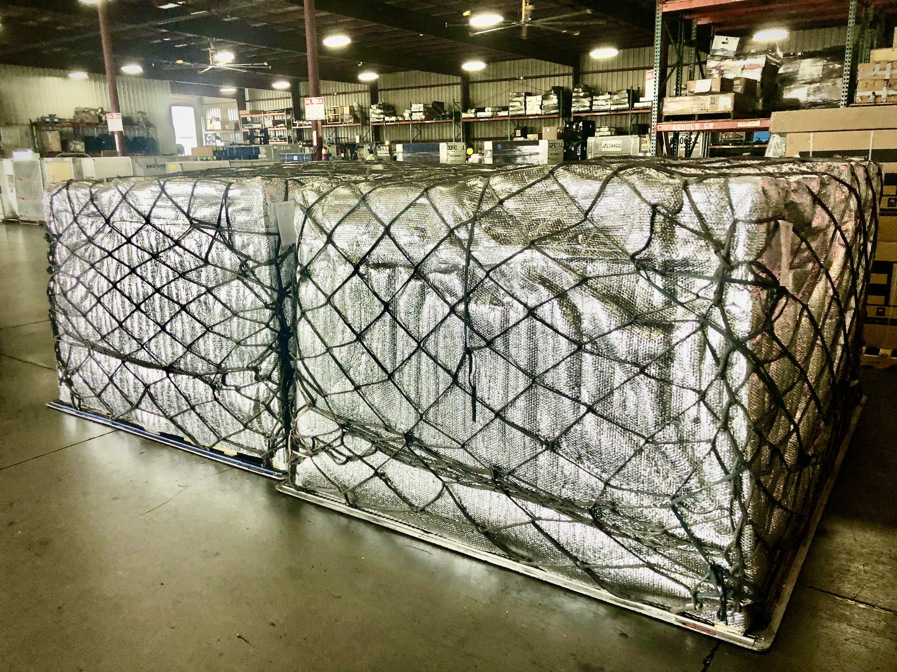 A large cargo aircraft operated by Pacific Air Cargo is seen on an airport tarmac with its nose lifted open to allow for front-loading of oversized freight. A massive wooden crate is being loaded into the aircraft using a heavy-duty scissor lift platform. Several ground crew members in high-visibility jackets are assisting with the operation, while others stand near a set of mobile stairs leading into the aircraft. The plane is a Boeing 747-400F, identifiable by its four engines and distinctive hump-shaped upper deck. The bright blue sky and modern terminal buildings in the background suggest a busy international airport setting. The scene captures the scale and precision of Pacific Air Cargo's logistics operations, highlighting their capability to handle oversized and high-value cargo.