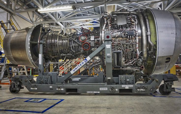 A large cargo aircraft operated by Pacific Air Cargo is seen on an airport tarmac with its nose lifted open to allow for front-loading of oversized freight. A massive wooden crate is being loaded into the aircraft using a heavy-duty scissor lift platform. Several ground crew members in high-visibility jackets are assisting with the operation, while others stand near a set of mobile stairs leading into the aircraft. The plane is a Boeing 747-400F, identifiable by its four engines and distinctive hump-shaped upper deck. The bright blue sky and modern terminal buildings in the background suggest a busy international airport setting. The scene captures the scale and precision of Pacific Air Cargo's logistics operations, highlighting their capability to handle oversized and high-value cargo.