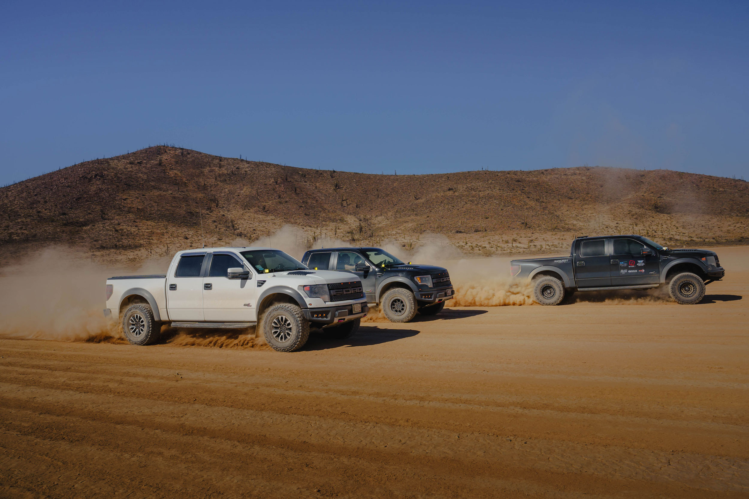 Three Ford Raptor trucks racing through desert terrain kicking up dust