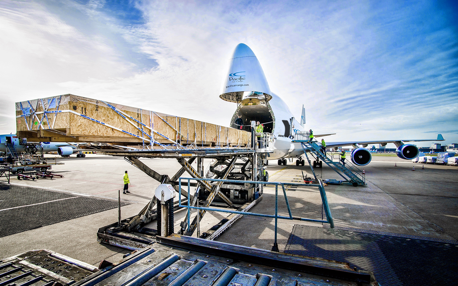 A Pacific Air Cargo Boeing 747 freighter parked on the tarmac with its nose cone raised open, while a long wooden crate is loaded through the front cargo door using a scissor-lift loader. Ground crew in high-visibility vests work on the loading platform un