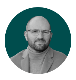 Professional headshot of a bald man with glasses and a beard, wearing a turtleneck and blazer, posed against a teal background.