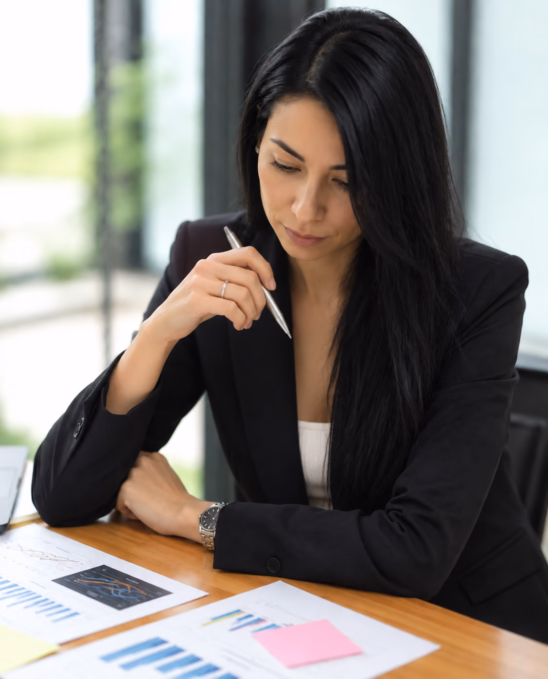Woman with long black hair in a black blazer sitting at a wooden table, holding a pen and looking thoughtfully at printed charts and graphs in front of her.