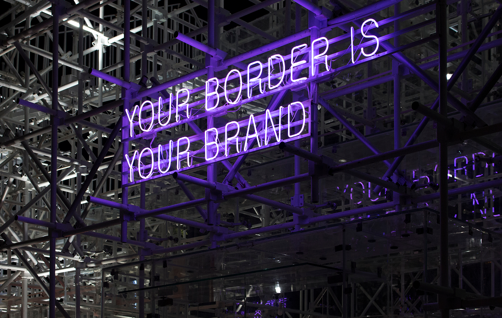 Purple neon sign reading 'YOUR BORDER IS YOUR BRAND' mounted on a complex metal scaffolding structure.