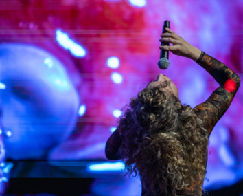 Curly-haired performer holding a microphone up while singing on stage with colorful blurred lights in the background.