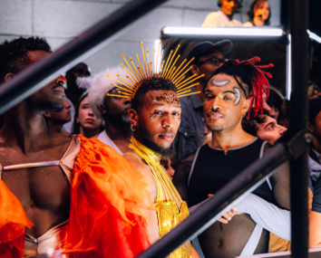 Group of fashionably dressed people, including a person wearing a golden crown-like headpiece and bright orange clothing.