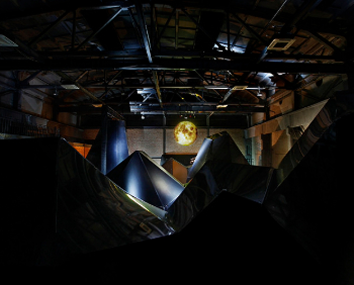 Dimly lit industrial space with abstract black angular sculptures and a glowing spherical light fixture hanging from the ceiling.