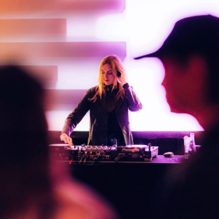 Female DJ performing at a club with colorful illuminated background panels.