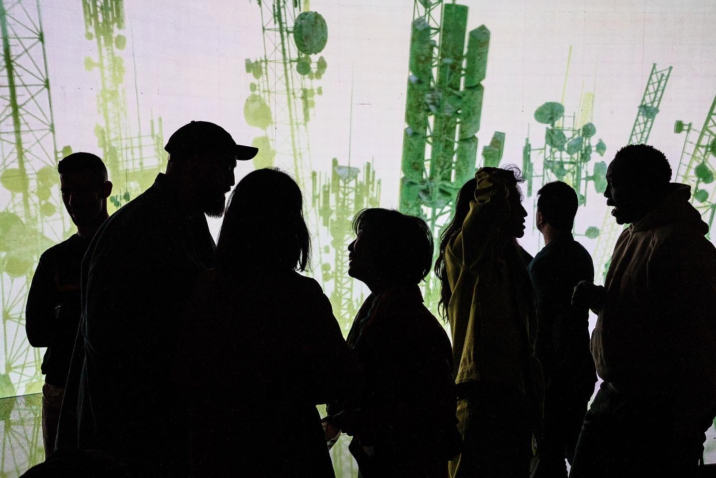 Silhouettes of seven people standing and interacting against a backdrop of green communication towers.