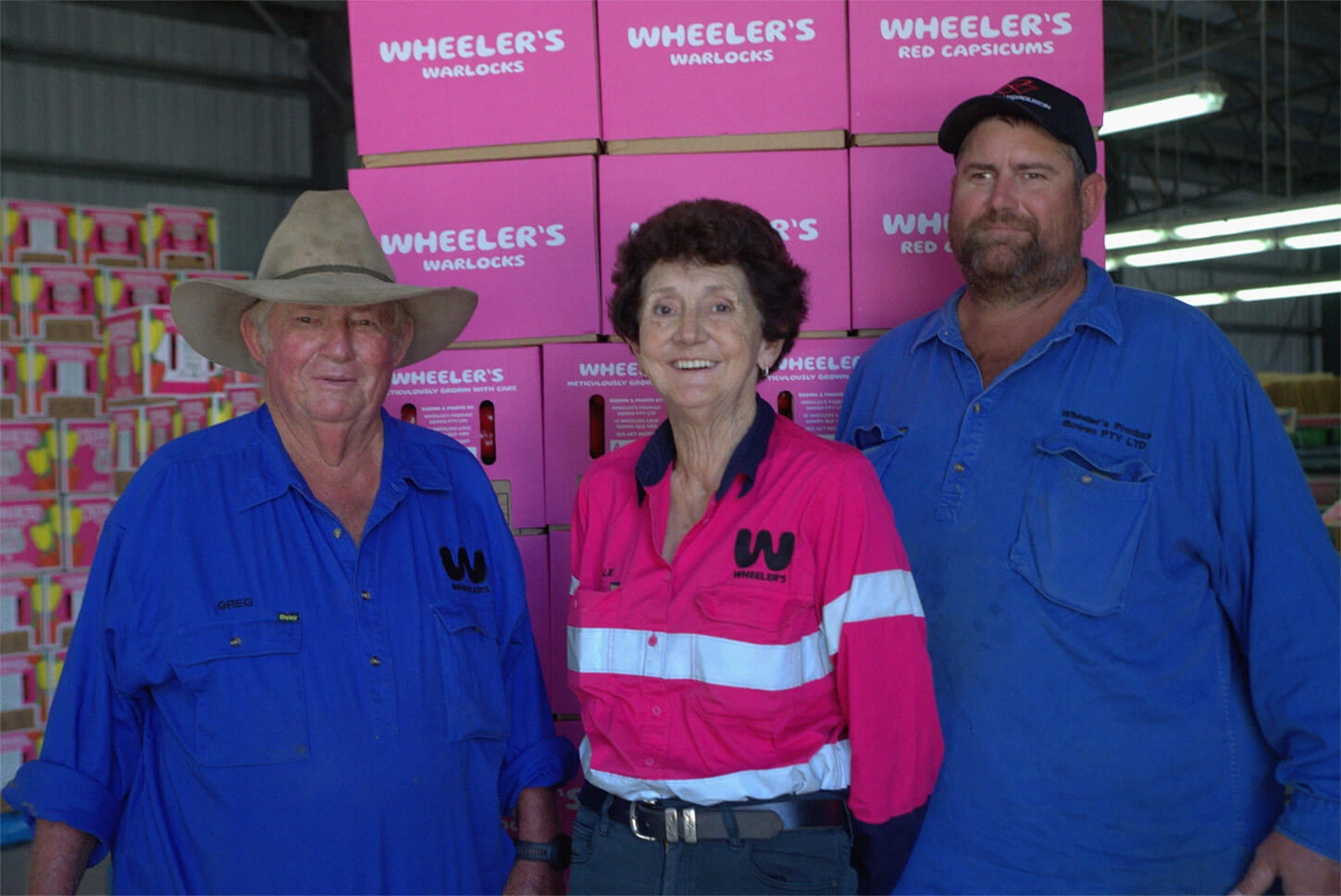 Greg, Natalie, and Tex — farmers from Wheeler’s Warlock — standing proudly beside a pallet stacked with pink produce boxes, each holding premium red Australian capsicums fresh from their farm.