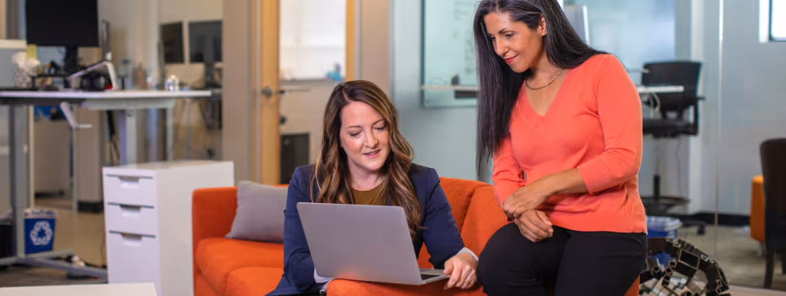 Two women founders in a casual modern office, working together on a laptop — representing collaborative firepower and strategic partnership