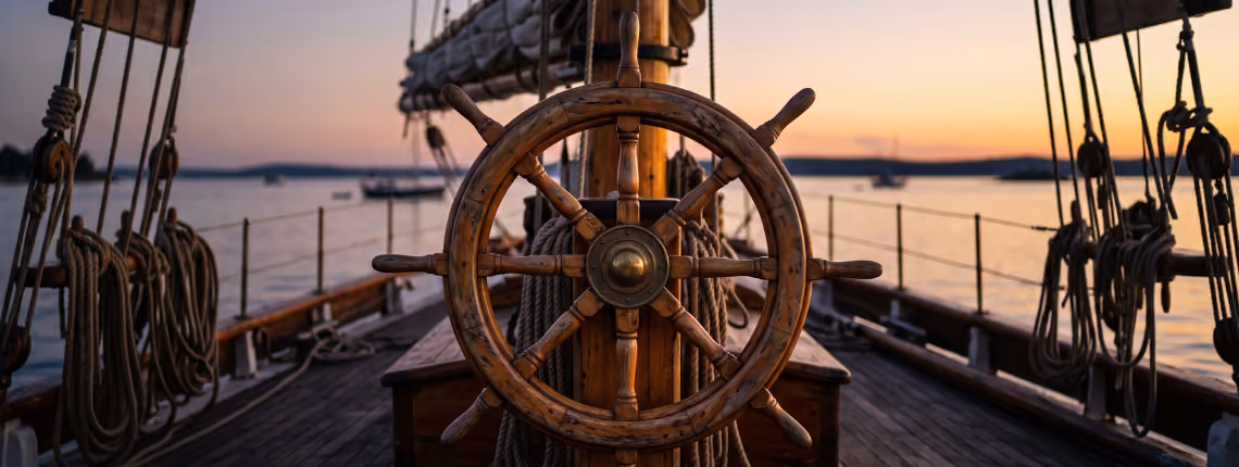 A weathered wooden ship's wheel on the deck of an old sailing boat at dusk, in warm evening light.