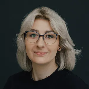 Porträt einer Frau mit blonden schulterlangen Haaren und einer Brille vor einem dunklen Hintergrund.