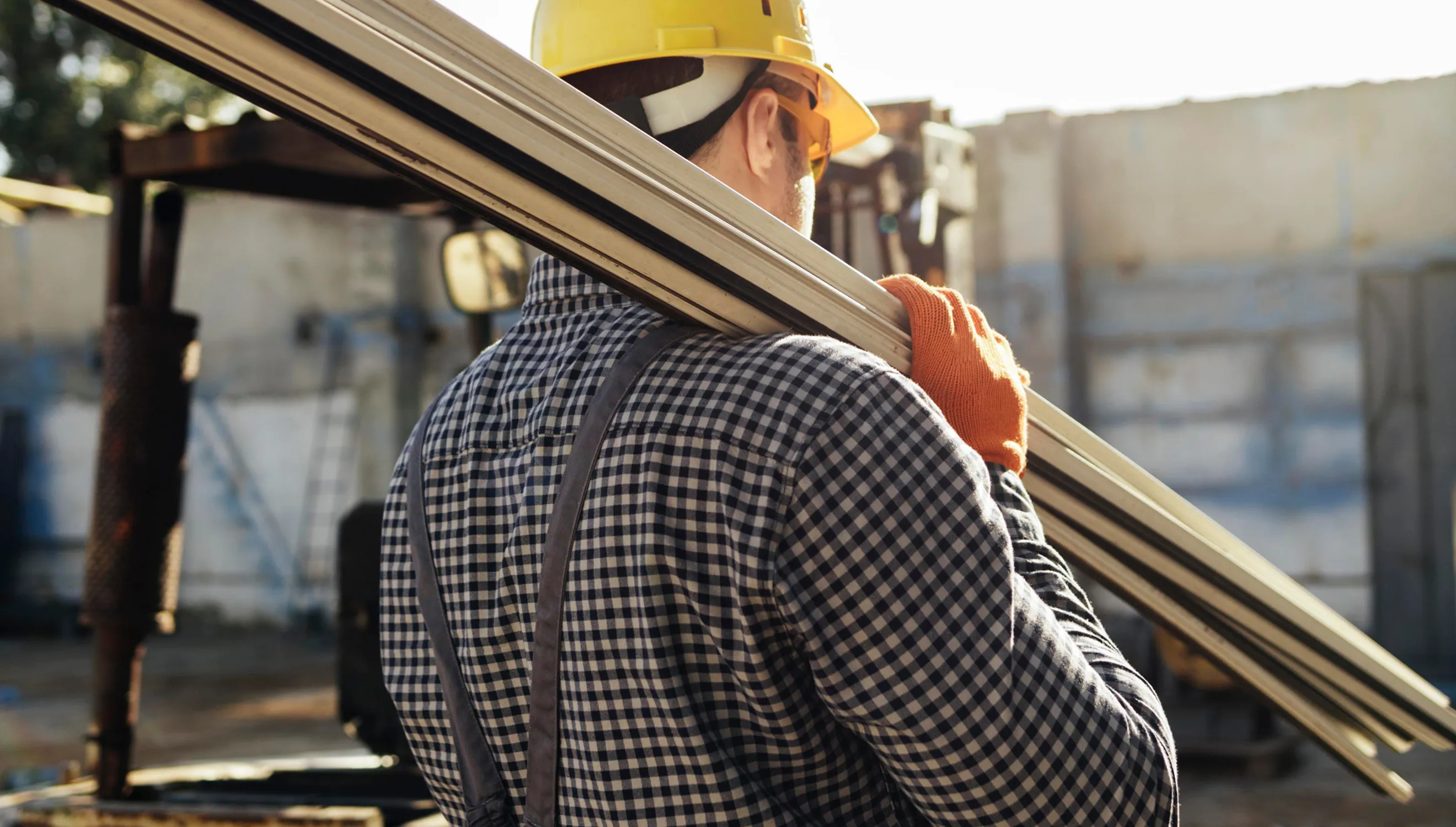 Ein Bauarbeiter mit gelbem Helm, orangenen Handschuhen und kariertem Hemd trägt mehrere lange Metallstangen auf der Schulter auf einer Baustelle.