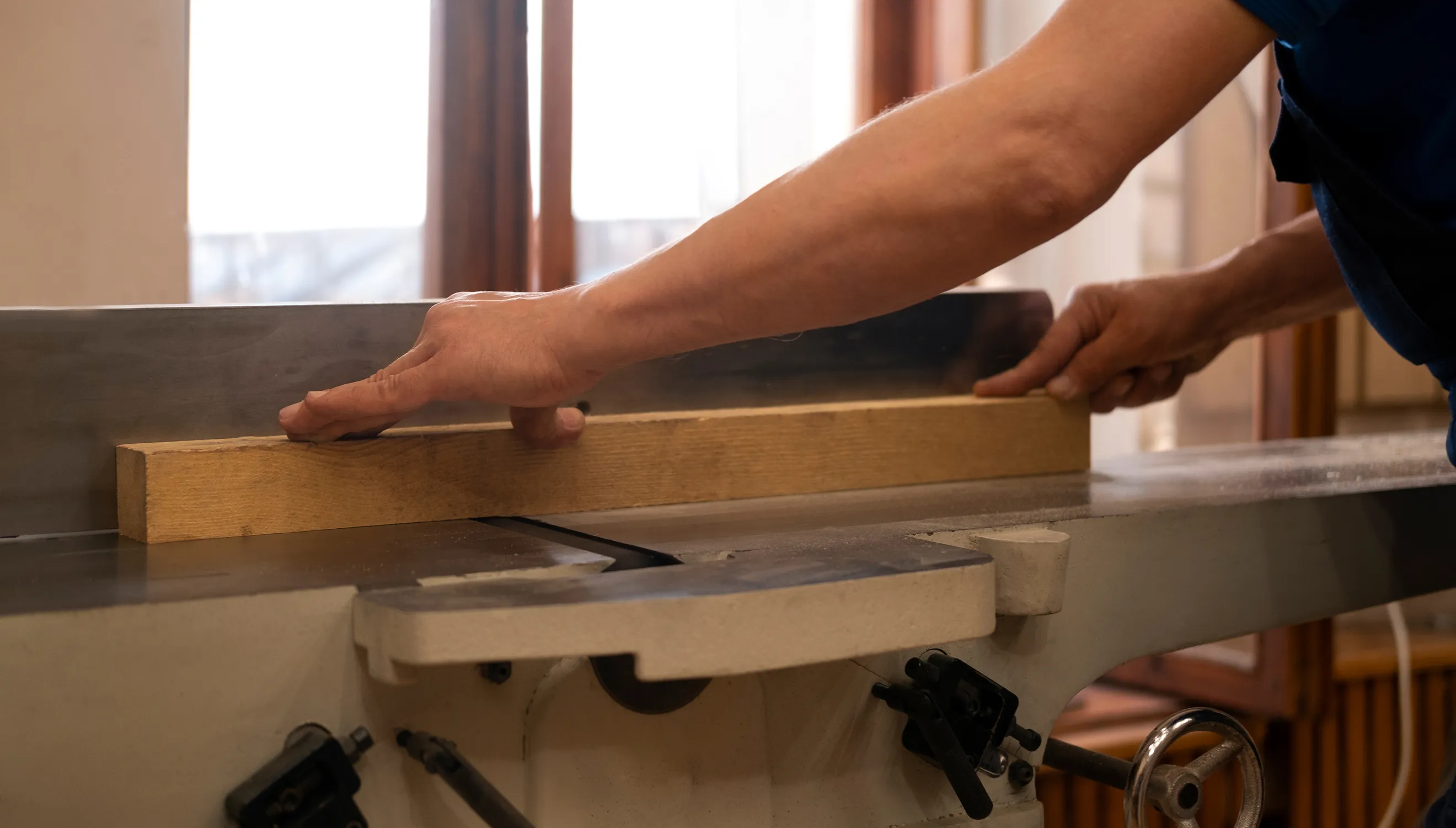 Person führt ein Stück Holz durch eine Hobelmaschine, um es zu glätten, mit beiden Händen am Holz auf einer Werkbank.