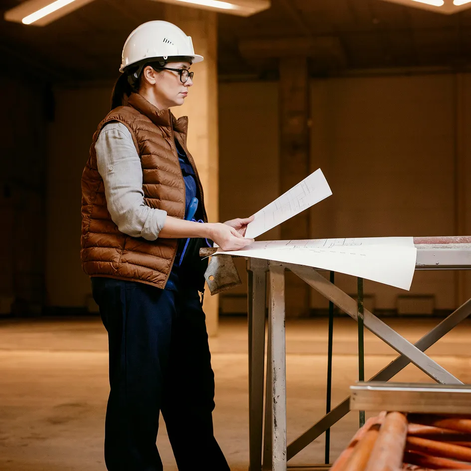 Frau auf einer Baustelle mit weißem Schutzhelm und brauner Weste, die Blaupausen auf einem Metalltisch überprüft.