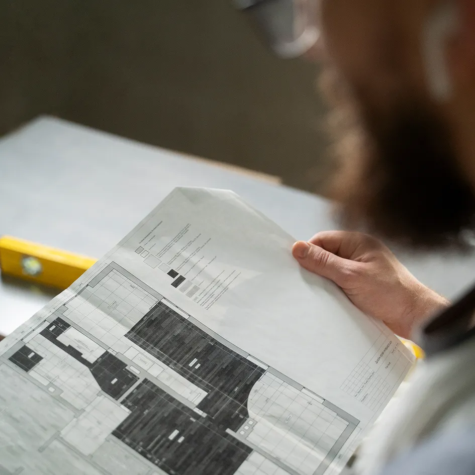 Person hält einen Architekturplan oder Bauzeichnung, im Hintergrund liegt eine Wasserwaage auf einem Tisch.
