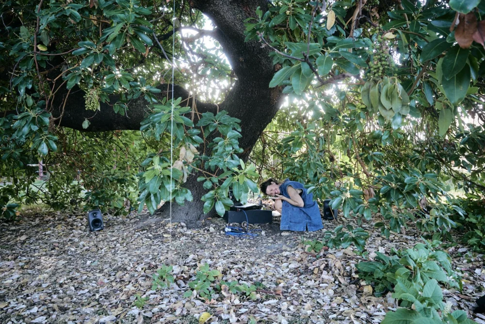 Maria sits under the tree next to her performance setup smiling 