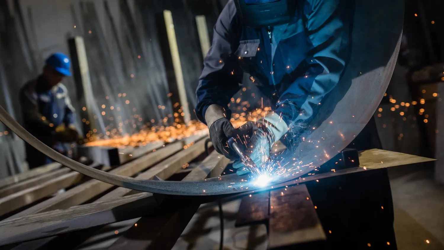 A welder in protective gear sparks as they work on a curved metal piece.