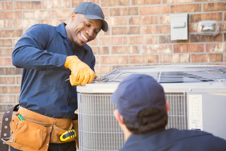 A smiling HVAC technician, who appears to be Black, is working on an air conditioner with a wrench.